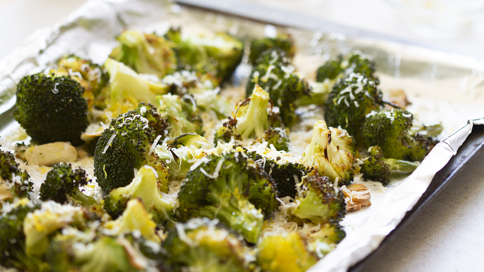 Roasted broccoli florets topped with grated cheese are spread on a foil-lined baking sheet. The vegetables are slightly browned and appear fresh out of the oven, with melted cheese sprinkled on top.