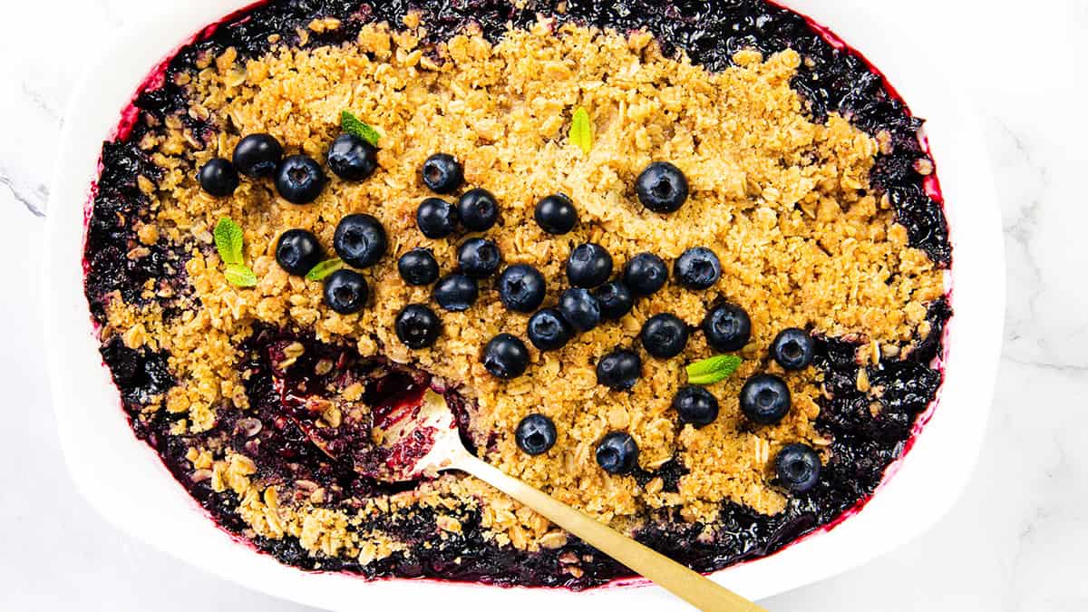 A baked blueberry crumble in a white dish, topped with fresh blueberries and mint leaves. Perfect for dessert recipes, it features a golden crumbly topping and juicy berry filling, served with a gold spoon for a tempting treat.
