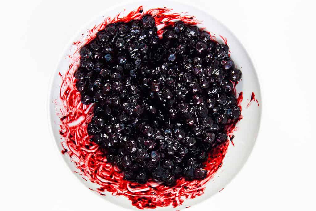 A white bowl filled with dark, juicy berries, likely blueberries, coated in a glossy red-purple syrup—perfect for a homemade Blueberry Crumble—with some of the syrup alluringly smeared along the sides of the bowl.