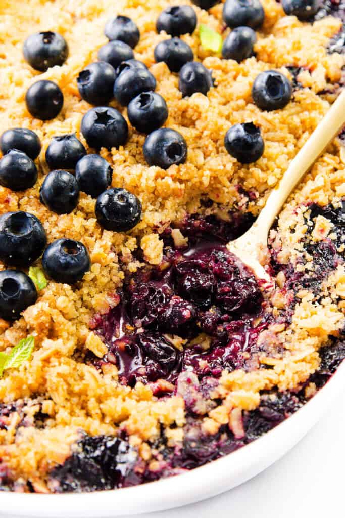 Close-up of a Blueberry Crumble in a white dish, topped with fresh blueberries and a golden crumb topping. A spoon scoops out a portion, revealing the juicy blueberry filling beneath this classic blueberry dessert.
