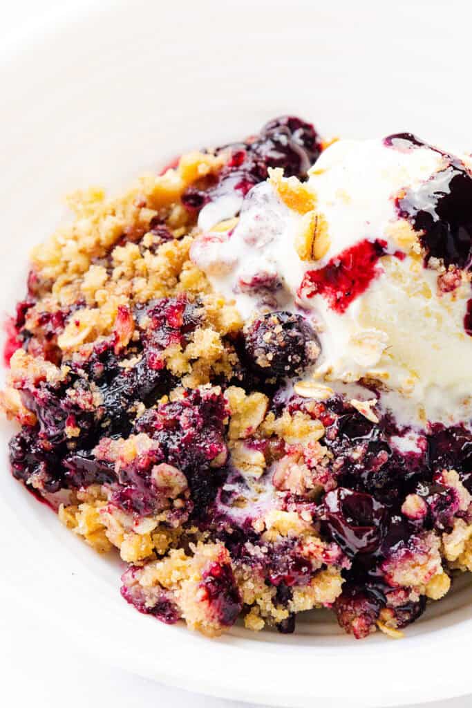 A close-up of a dessert bowl with blueberry crumble, topped with a scoop of vanilla ice cream. The crumble is golden and crunchy, with dark purple berries and melted ice cream blending on top.