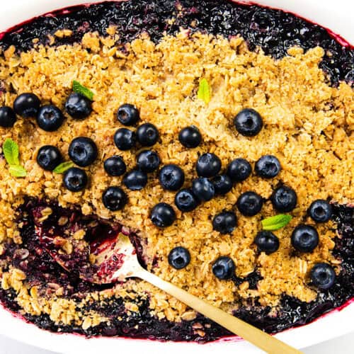 A baked blueberry crisp in a white dish, this delicious blueberry dessert features a golden crumbly streusel, fresh blueberries, and mint leaves; a spoon rests in the dish where a portion of the fruit crumble has been served.
