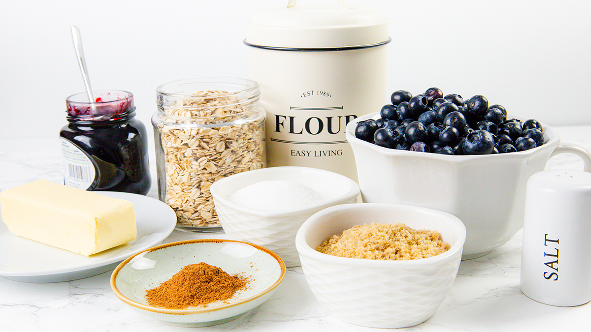A collection of baking ingredients on a white surface, including butter, cinnamon, oats, brown and white sugar, flour, blueberries in a bowl, salt, and a jar of jam.