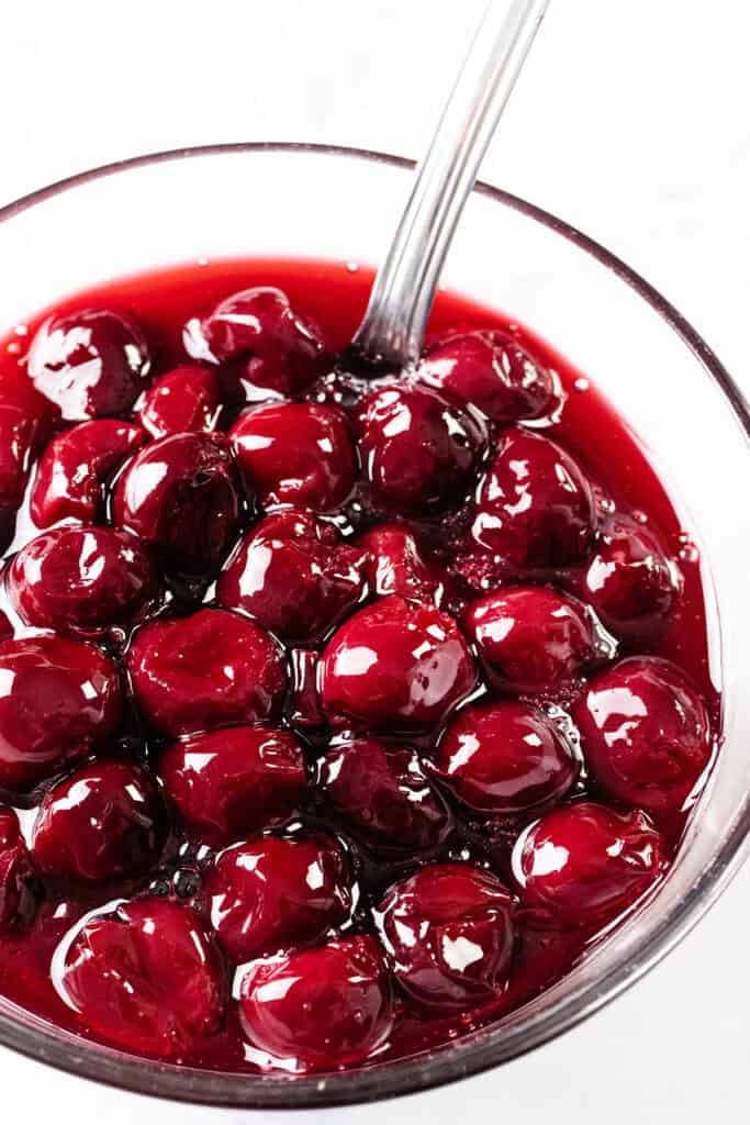 A close-up of a glass bowl filled with glossy, red cherries in syrup, with a metal spoon resting inside—perfect for preparing a classic Cherries Jubilee recipe or enjoying as part of a flaming dessert.