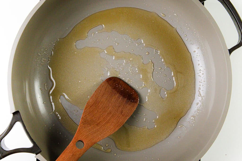 A gray frying pan with a light coating of oil being spread by a wooden spatula, ready to start a classic Flaming Cherries Jubilee dessert. The pan sits on a white surface, and the oil glistens under the light.