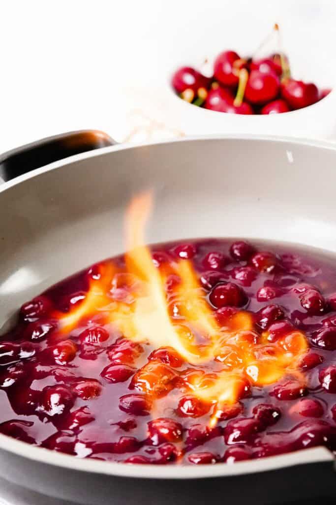 A close-up of a skillet with Flaming Cherries Jubilee sauce, showing whole cherries in a glossy red liquid. In the background, a bowl of fresh cherries is slightly out of focus, capturing the flair of this classic flaming dessert.