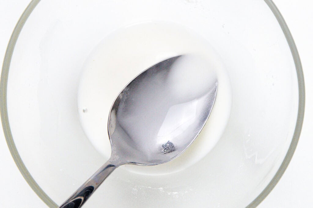A metal spoon rests in a glass bowl containing a small amount of white liquid, likely a mixture such as icing or glaze, reminiscent of preparations for a classic flaming dessert like Cherries Jubilee, on a white background.