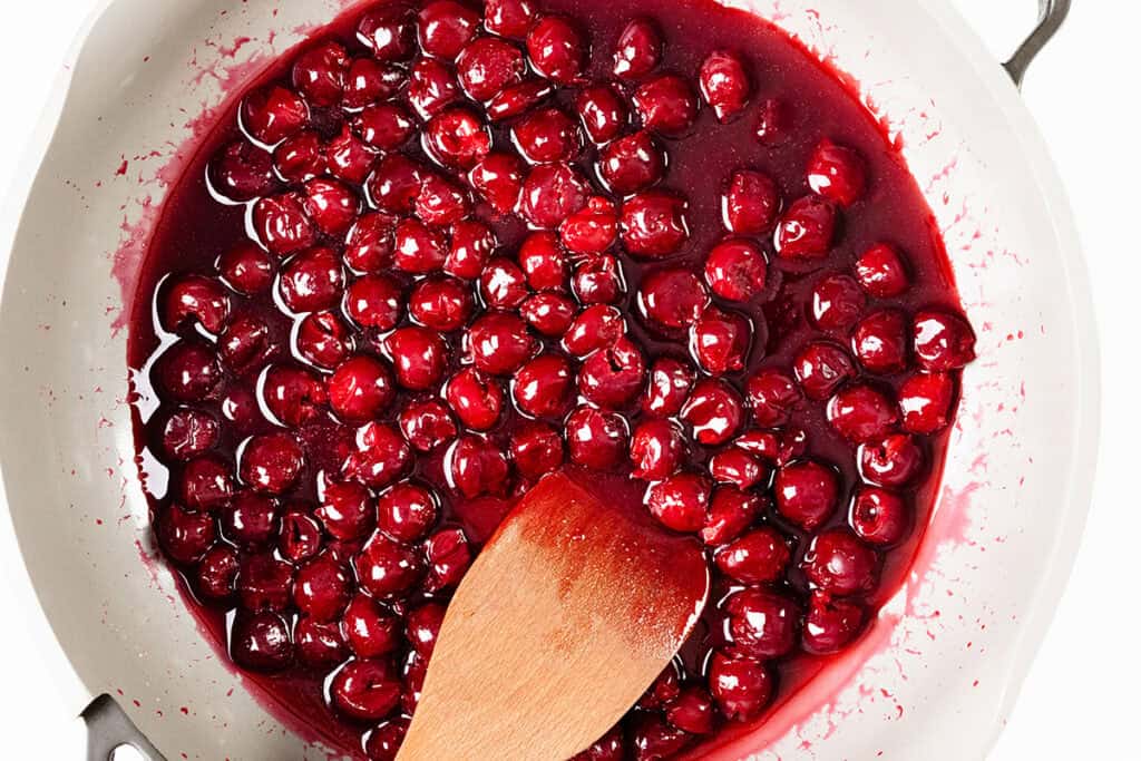 A wooden spoon stirs glossy, cooked cherries in a thick red sauce inside a white pan—perfect for creating a classic Flaming Cherries Jubilee.