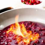 A close-up of Flaming Cherries Jubilee in a pan, cherries in syrup being flambéed with a vibrant orange flame. In the background, a bowl of fresh cherries sits slightly out of focus, highlighting this classic flaming dessert.