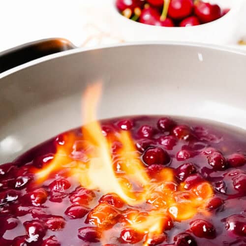 A close-up of Flaming Cherries Jubilee in a pan, cherries in syrup being flambéed with a vibrant orange flame. In the background, a bowl of fresh cherries sits slightly out of focus, highlighting this classic flaming dessert.