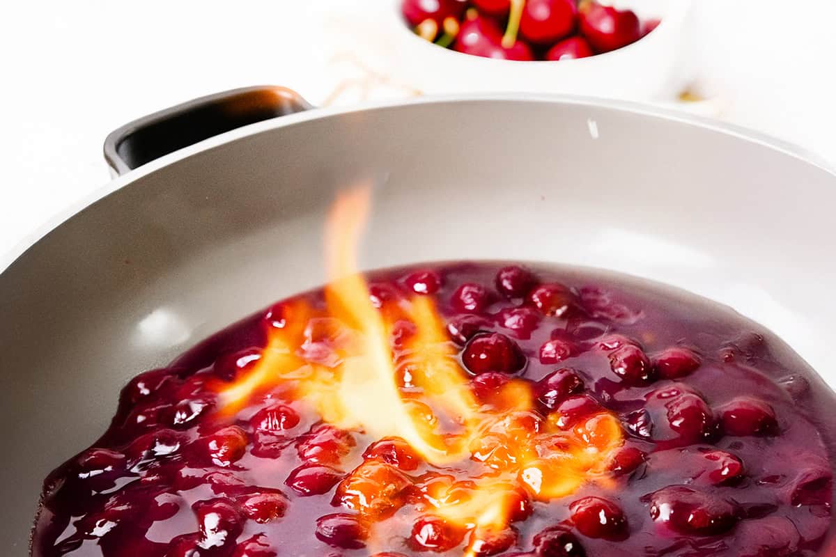 A close-up of Flaming Cherries Jubilee in a pan, cherries in syrup being flambéed with a vibrant orange flame. In the background, a bowl of fresh cherries sits slightly out of focus, highlighting this classic flaming dessert.