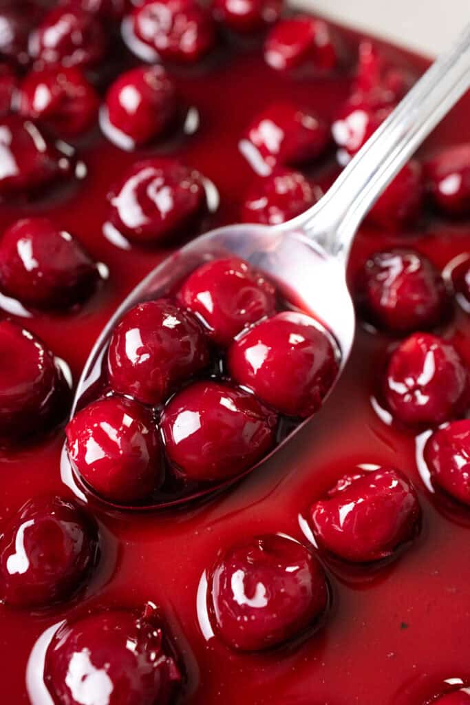 A close-up of glossy, bright red cherries in syrup, with a metal spoon scooping up several cherries from the mixture—perfect for creating Flaming Cherries Jubilee, a classic flaming dessert.