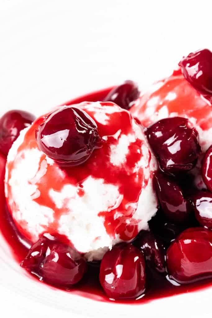 Two scoops of vanilla ice cream topped with glossy, red cherry sauce and whole cherries, served on a white plate for a delightful take on the classic Flaming Cherries Jubilee dessert.