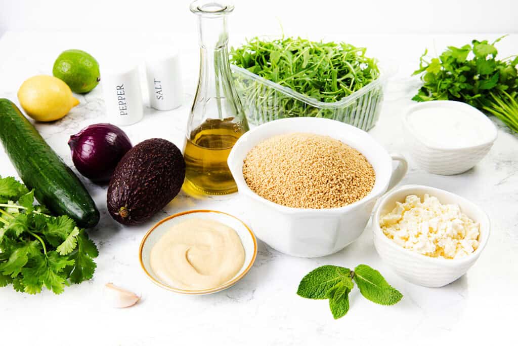 A variety of fresh ingredients and seasonings on a white surface, perfect for a couscous salad—featuring cucumber, red onion, avocado, lemon, lime, garlic, herbs, greens, grains, feta cheese, oil, tahini, yogurt, salt and pepper.