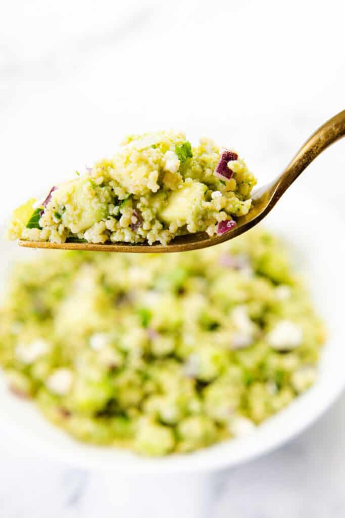 A close-up of a fork holding a bite of couscous salad with green herbs, red onion, and avocado, tossed in zesty Lemon Lime Dressing, with a blurred bowl of the same salad in the background.
