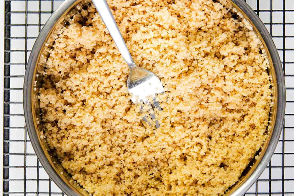 A metal bowl filled with cooked couscous sits on a wire rack. A silver fork is placed inside the bowl, its tines buried in the fluffy, golden grains—perfect for making a vibrant couscous salad with Lemon Lime Dressing.