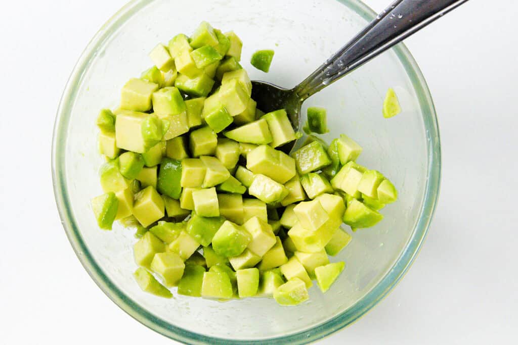 A glass bowl filled with diced avocado pieces, perfect for adding to a couscous salad. A silver spoon rests inside, and the evenly chopped green avocado sits on a white surface.