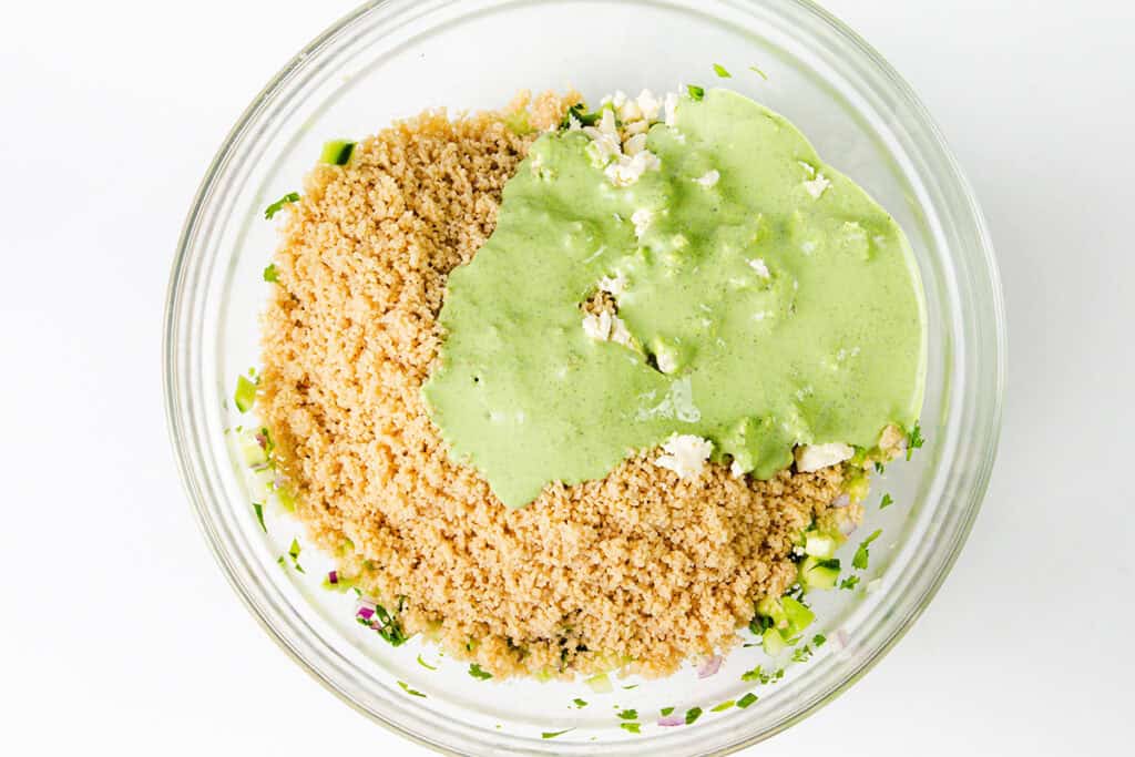 A glass bowl contains breadcrumbs, chopped herbs, and a creamy light green cilantro dressing, partially mixed together, on a white background.