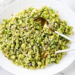 A large white plate holds a green, finely chopped Couscous Salad with vegetables and grains. Two serving utensils, one gold and one white, rest on the plate. The background is a marble surface with part of a white cloth visible.