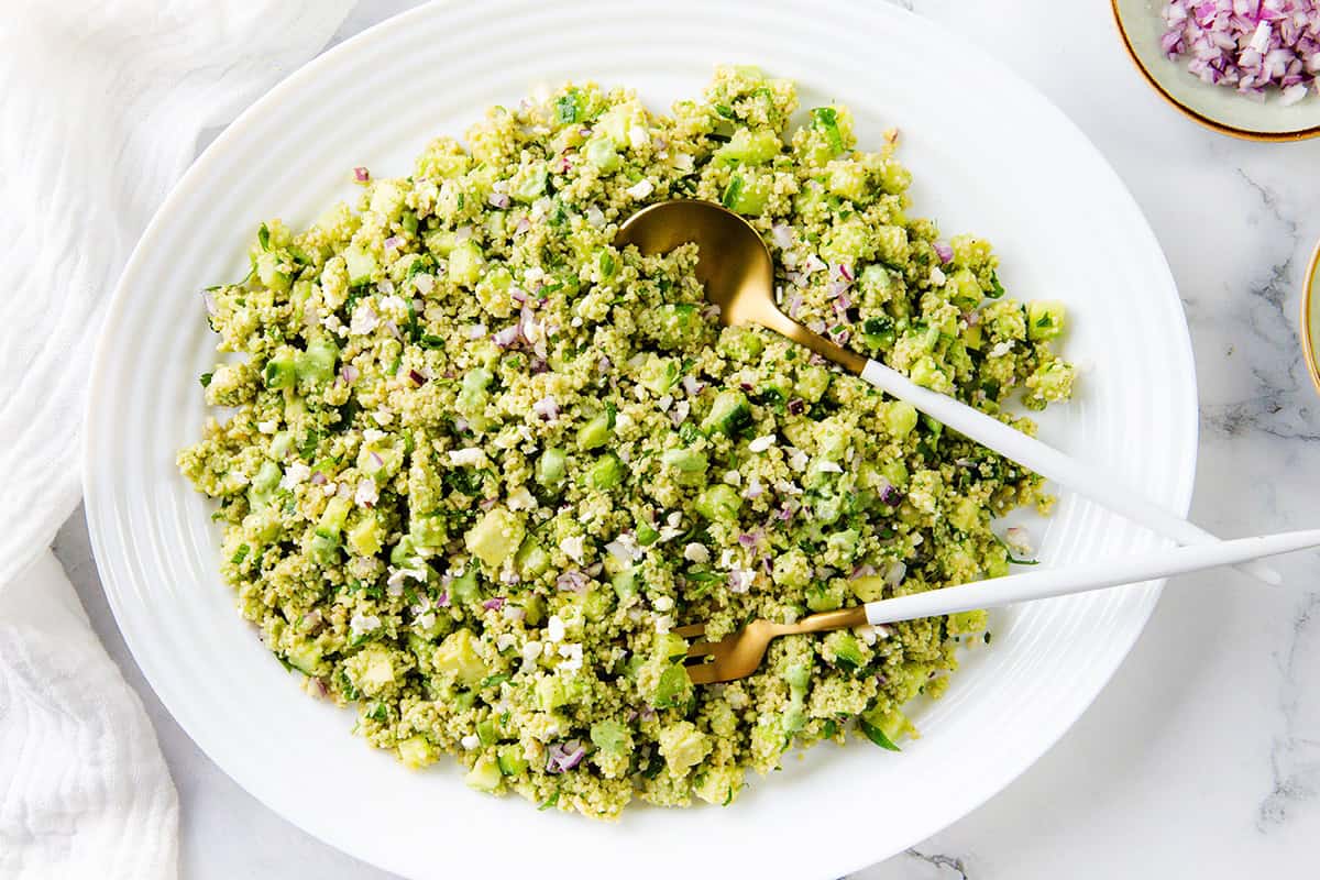 A large white plate holds a green, finely chopped Couscous Salad with vegetables and grains. Two serving utensils, one gold and one white, rest on the plate. The background is a marble surface with part of a white cloth visible.