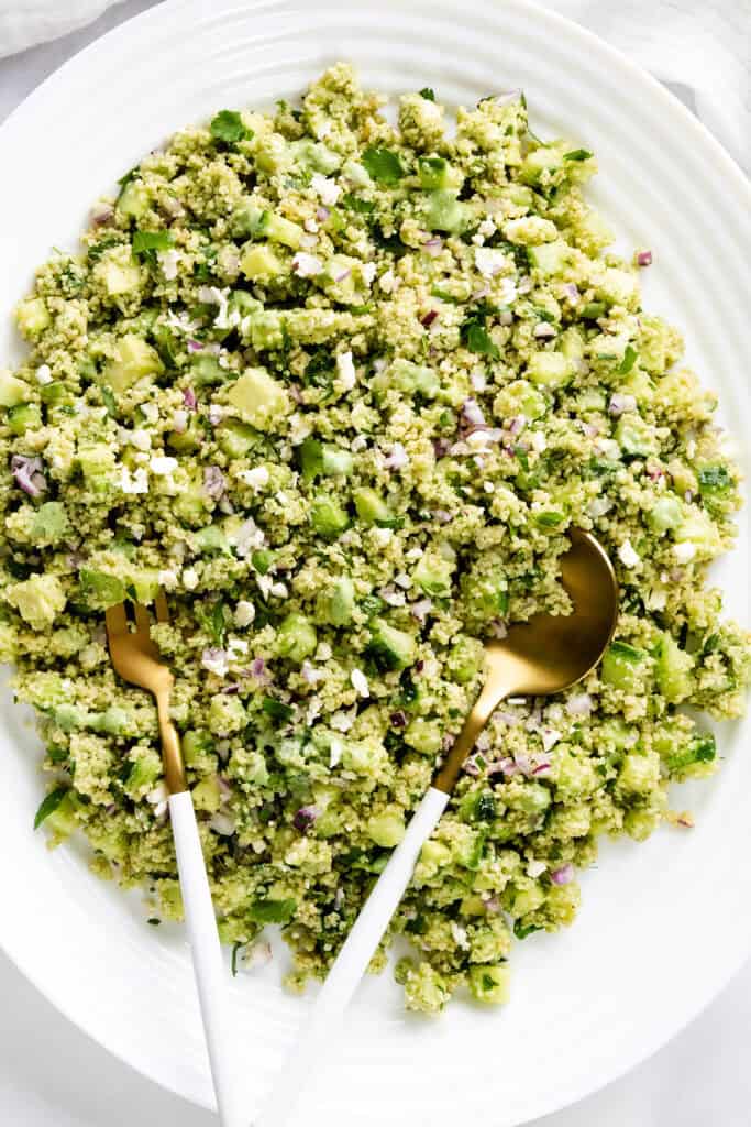A large white plate filled with a green couscous salad, garnished with chopped herbs and vegetables, topped with a zesty lemon lime dressing. A gold fork and spoon are placed on top.