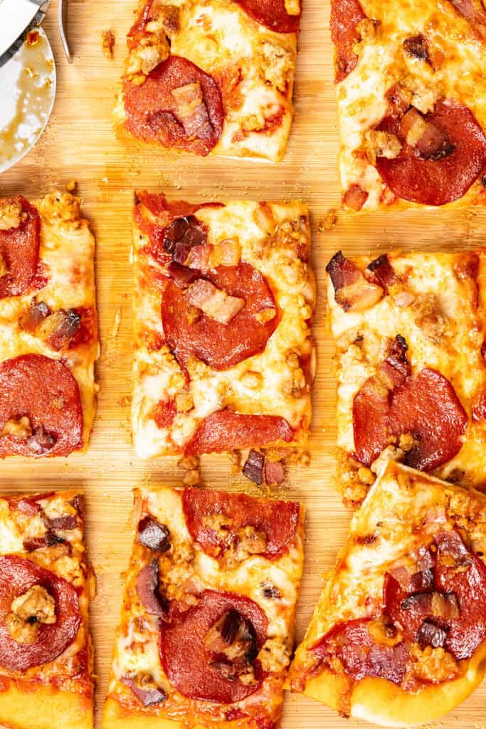 Rectangular slices of Meat Lovers Pizza with pepperoni, sausage, and bacon rest on a wooden cutting board. Some slices are separated, revealing a golden, cheesy crust and browned toppings. A pizza cutter sits at the edge.