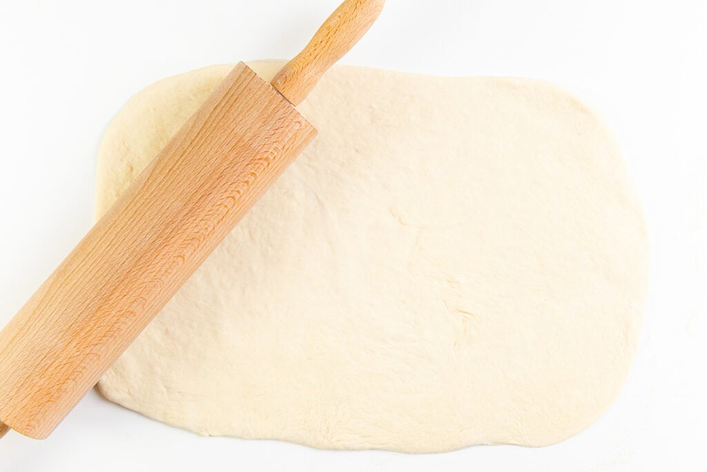 A wooden rolling pin rests on top of a sheet of rolled-out dough on a white surface, ready to be transformed into a delicious Meat Lovers Pizza.