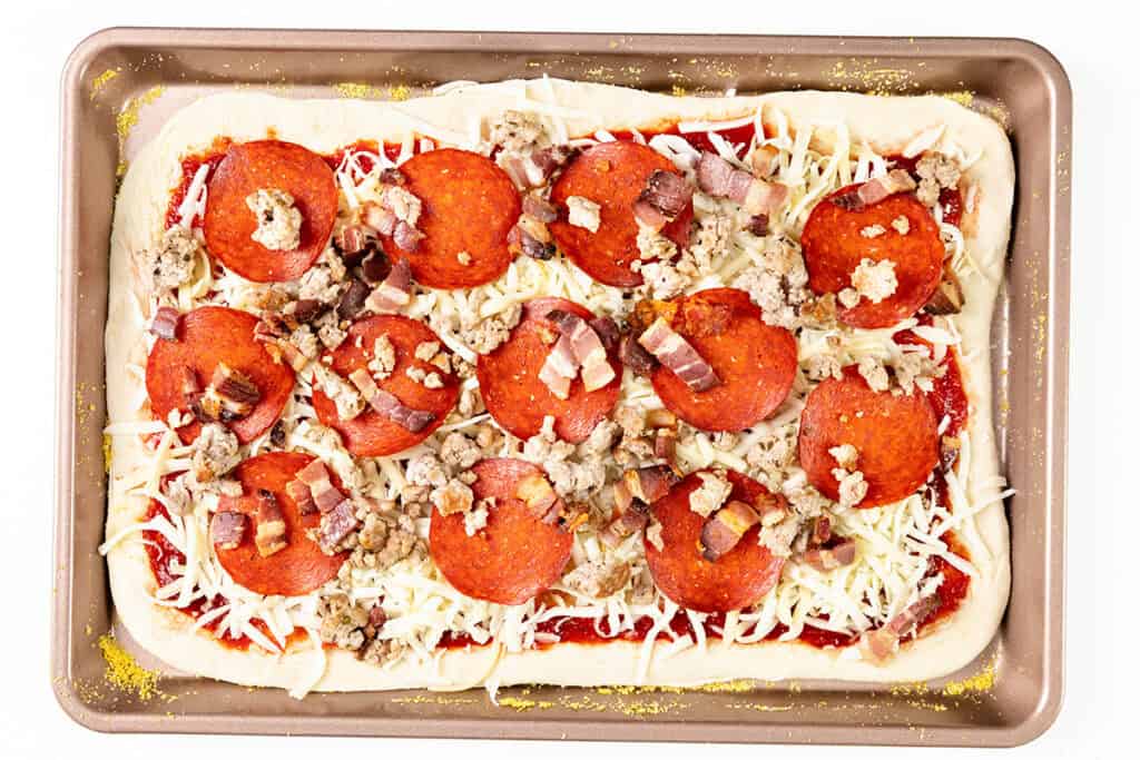 Rectangular homemade Meat Lovers Pizza on a baking sheet, topped with mozzarella cheese, pepperoni slices, sausage, bacon pieces, and tomato sauce, with a hint of cornmeal scattered along the pan edges.