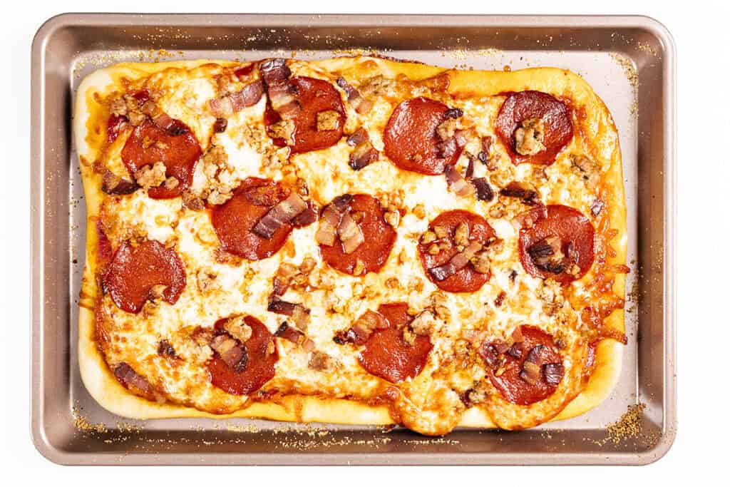 Rectangular homemade Meat Lovers Pizza on a baking sheet, topped with melted cheese, pepperoni slices, crumbled sausage, and pieces of bacon. The golden brown crust and bubbly edges make this pizza a must for any meat lover.