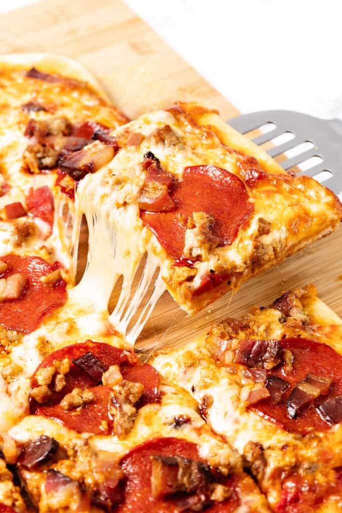 A metal spatula lifts a cheesy slice of rectangular Meat Lovers Pizza, stretching melted cheese, from a wooden cutting board. The pizza features pepperoni and crumbled sausage pieces.