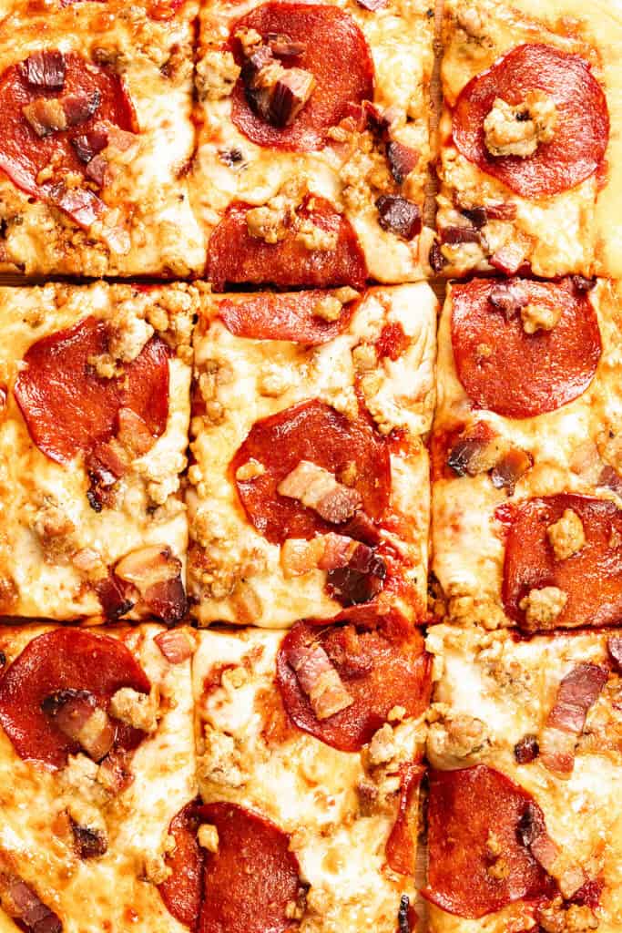 Close-up of a Meat Lovers Pizza cut into nine square slices, topped with melted cheese, pepperoni, crumbled sausage, and small pieces of bacon.