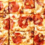 Close-up of a Meat Lovers Pizza cut into square slices, topped with melted cheese, pepperoni, sausage, and pieces of bacon. The toppings are evenly distributed across the golden-brown surface.