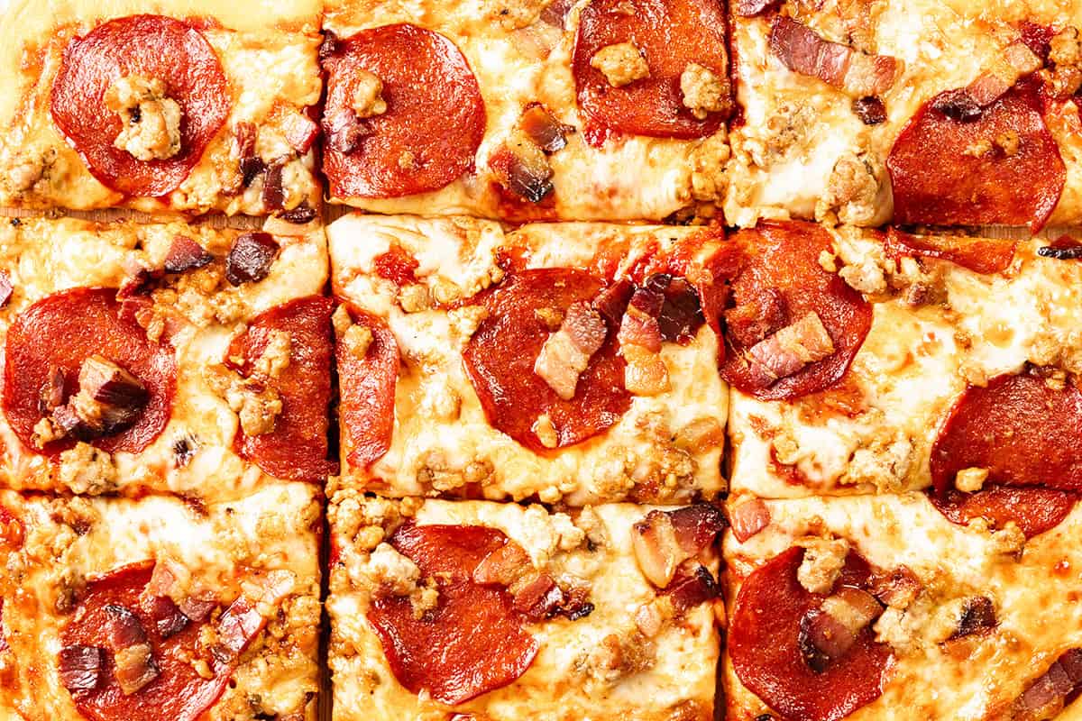 Close-up of a Meat Lovers Pizza cut into square slices, topped with melted cheese, pepperoni, sausage, and pieces of bacon. The toppings are evenly distributed across the golden-brown surface.