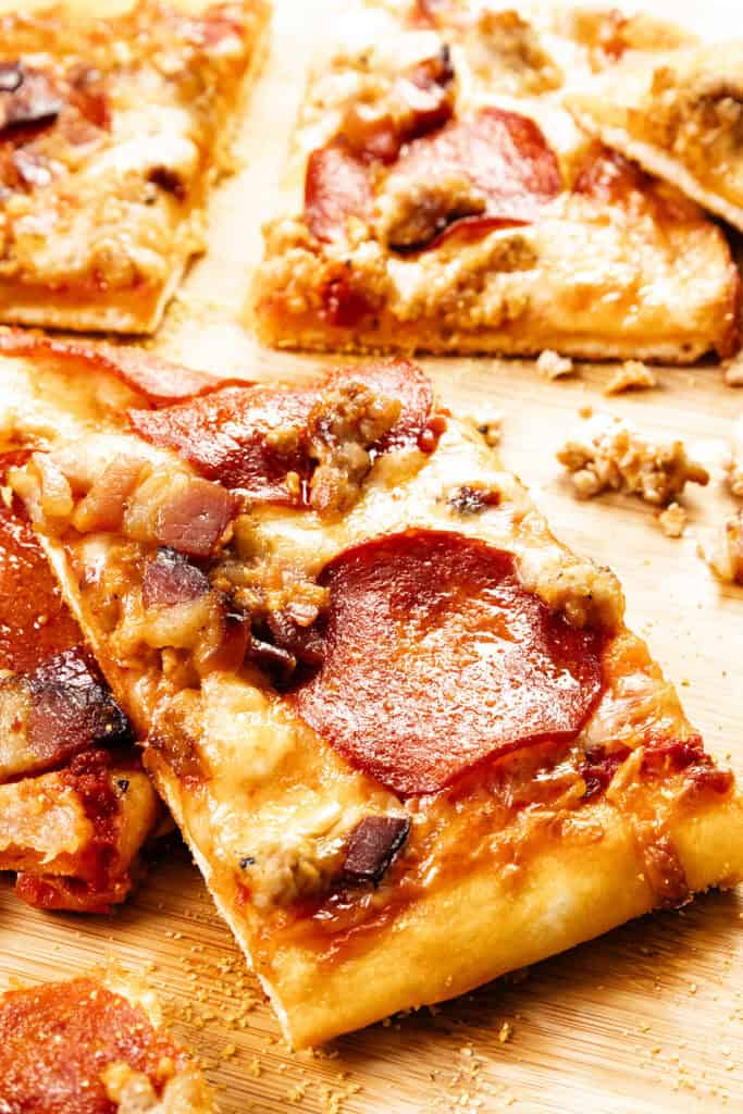 Close-up of a rectangular slice of Meat Lovers Pizza topped with pepperoni, sausage, bacon, and melted cheese on a wooden surface, with other pizza slices visible in the background.