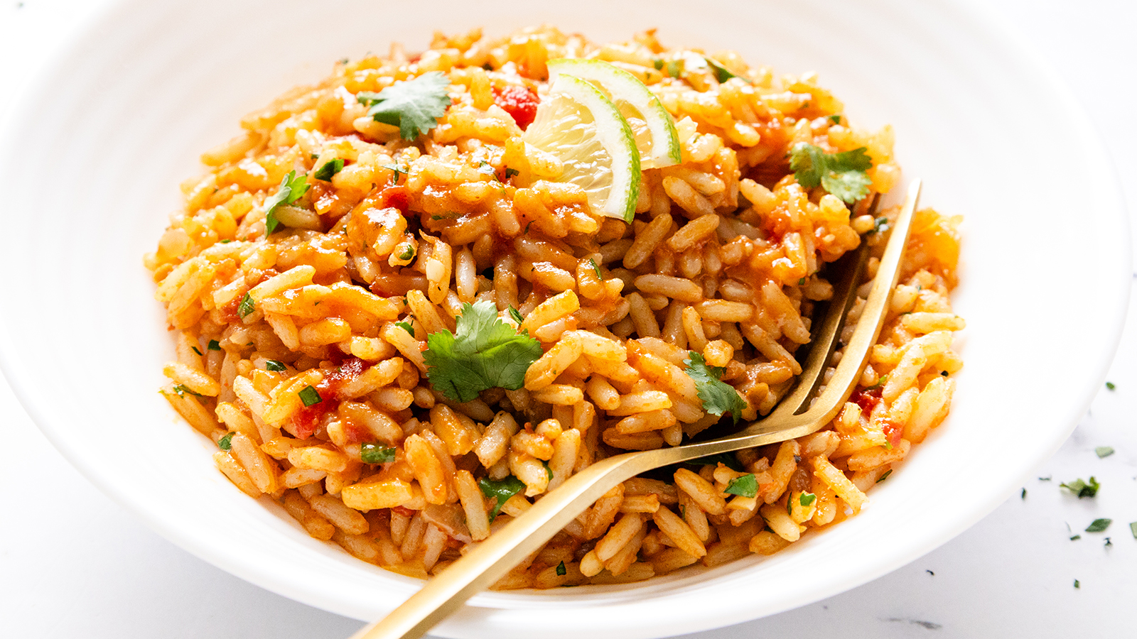 A bowl of seasoned rice with visible herbs, tomato sauce, and garnished with fresh cilantro and lime wedges, served with a gold fork.
