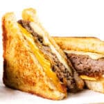 Two halves of a classic diner-style patty melt, featuring melted cheddar and Swiss cheese, a juicy beef patty, and caramelized onions on toasted bread, served with a small metal cup of onions on the side.