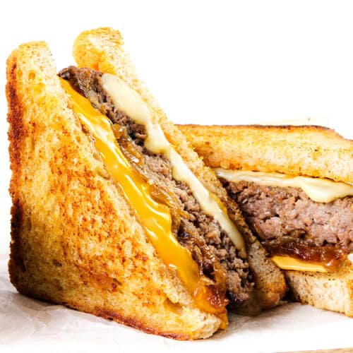 Two halves of a classic diner-style patty melt, featuring melted cheddar and Swiss cheese, a juicy beef patty, and caramelized onions on toasted bread, served with a small metal cup of onions on the side.