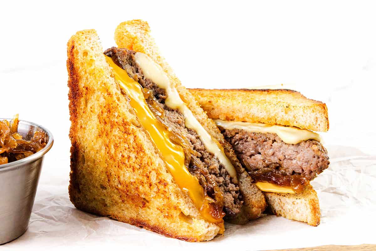 Two halves of a classic diner-style patty melt, featuring melted cheddar and Swiss cheese, a juicy beef patty, and caramelized onions on toasted bread, served with a small metal cup of onions on the side.
