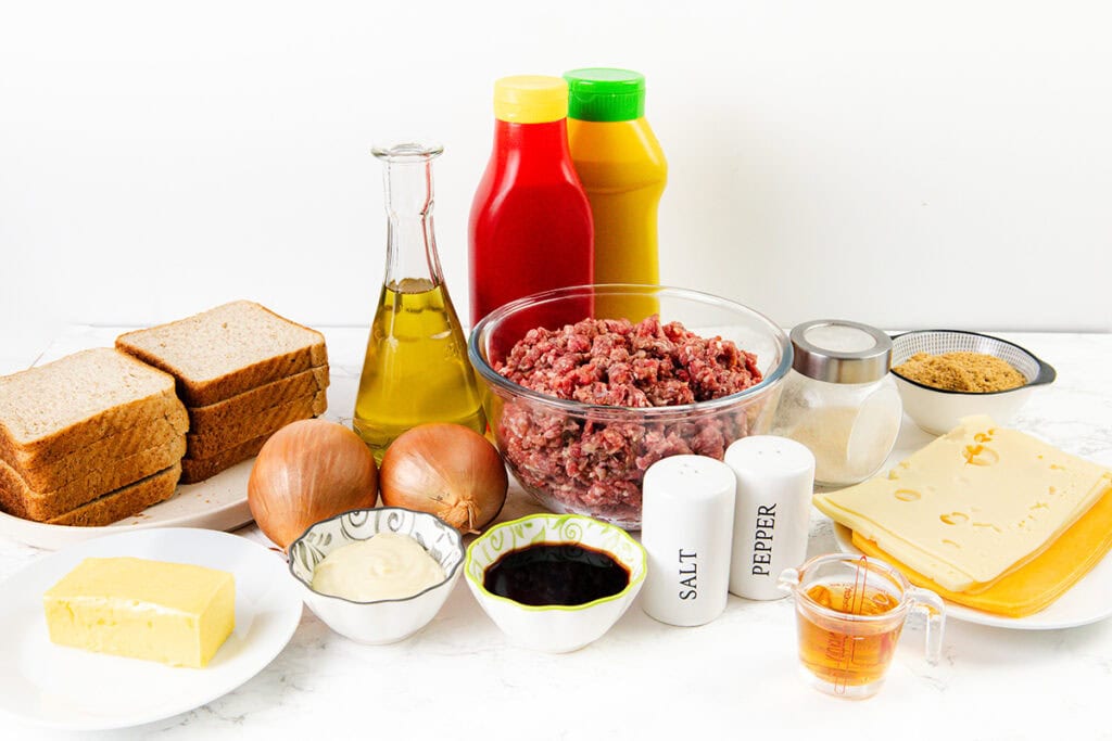 A variety of ingredients on a white surface, including sliced bread, raw ground beef in a glass bowl, cheese slices, onions, butter, oil, condiments, and seasonings—everything you need for a classic diner-style patty melt.