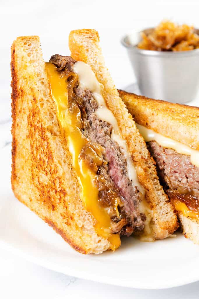 A close-up of a diner-style patty melt featuring melted cheddar, another cheese, caramelized onions, and a beef patty on toasted bread, served on a white plate. A small bowl of caramelized onions sits in the background.