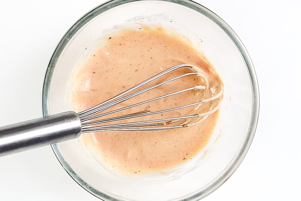 A metal whisk rests in a glass bowl filled with a creamy, light pink sauce or dressing—perfect for adding a classic, diner-style touch to your next Patty Melt—set against a white background.