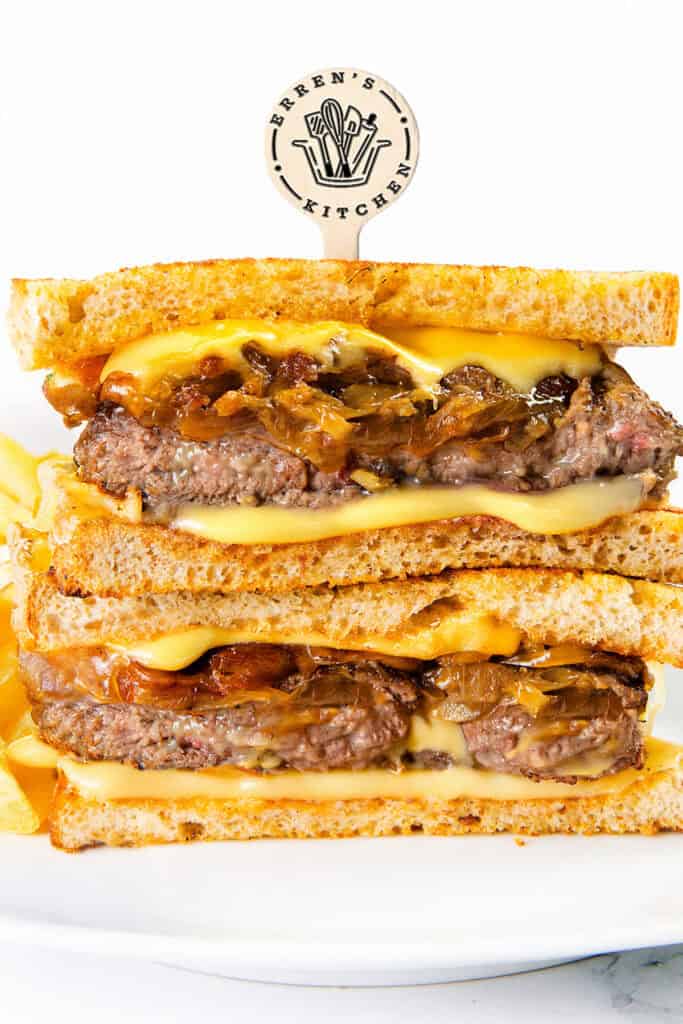 A close-up of a Patty Melt stacked sandwich with two layers of beef patties, melted cheese, and caramelized onions between toasted bread, held together with an "Erren's Kitchen" stick. Fries are visible on the side.