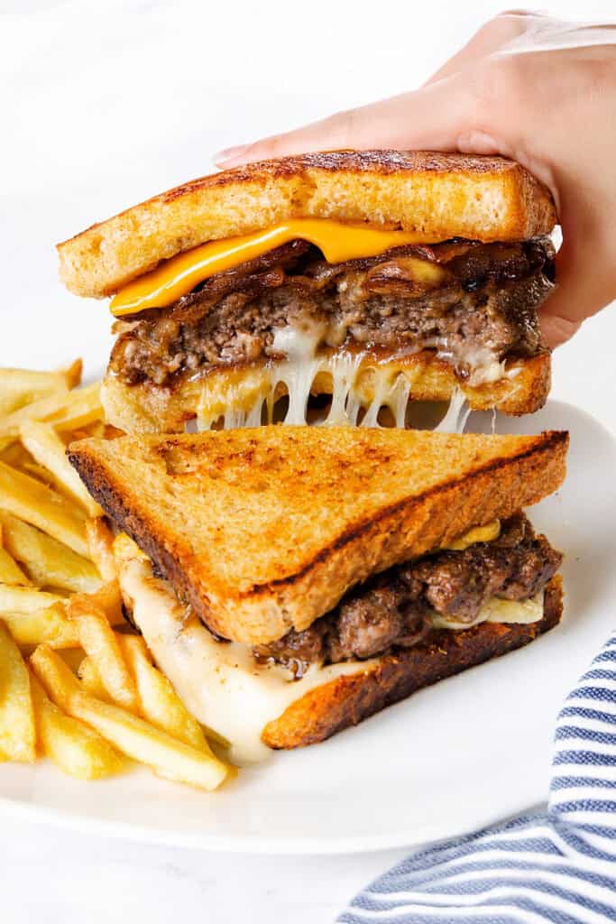 A hand holds half of a diner-style patty melt with melted cheese, beef patty, and bacon, above another half on a plate. French fries are served on the side.