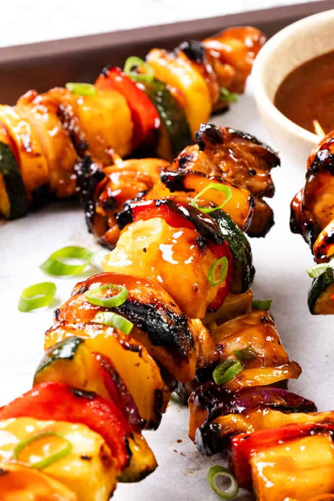 Close-up of BBQ chicken skewers with chunks of chicken, bell peppers, zucchini, and pineapple, glazed with sauce and garnished with sliced green onions. A small bowl of dipping sauce is visible in the background.
