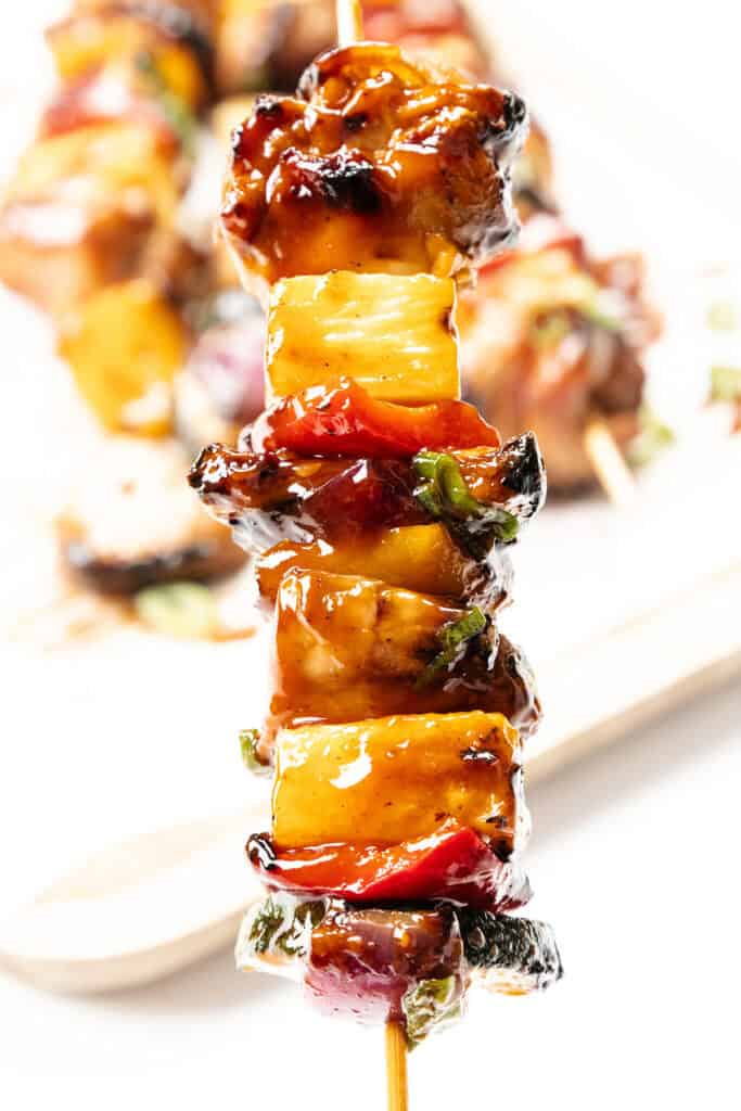 A close-up of Pineapple BBQ chicken skewers with glazed chunks of meat, pineapple, and colorful bell peppers, garnished with herbs. The background is blurred, showing more chicken skewers on a white plate.