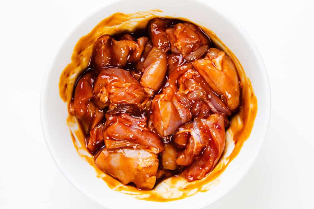 A white bowl filled with raw chicken pieces coated in a reddish-brown marinade, perfect for making BBQ skewers, set against a plain white background.