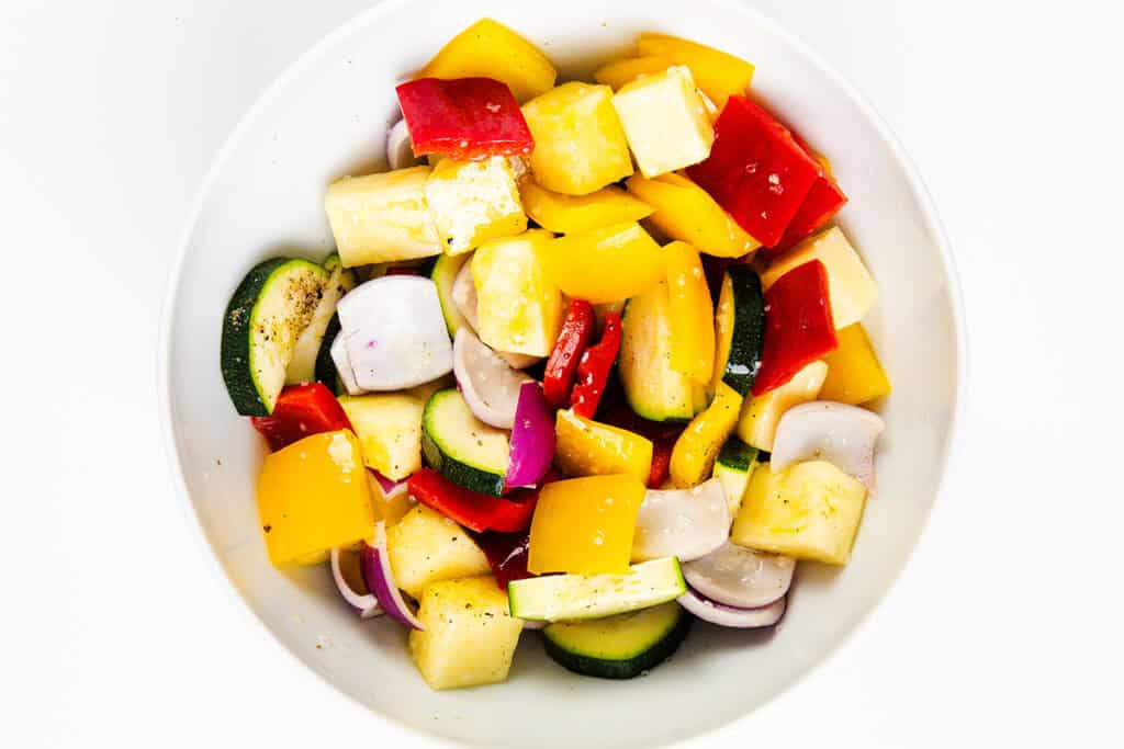 A white bowl filled with chopped colorful vegetables, including yellow and red bell peppers, zucchini, red onion, pineapple chunks, and pieces of yellow squash—perfect for threading onto skewers for your next BBQ.