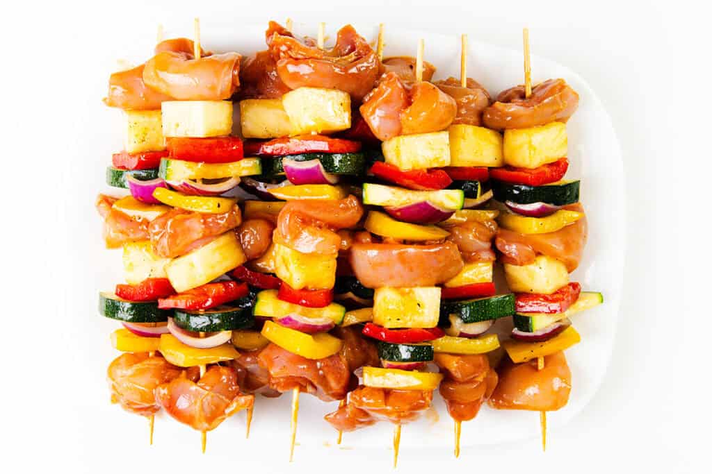 A plate of uncooked BBQ chicken skewers with marinated chicken, zucchini, red onion, yellow bell pepper, and pineapple chunks arranged neatly on wooden sticks against a white background.