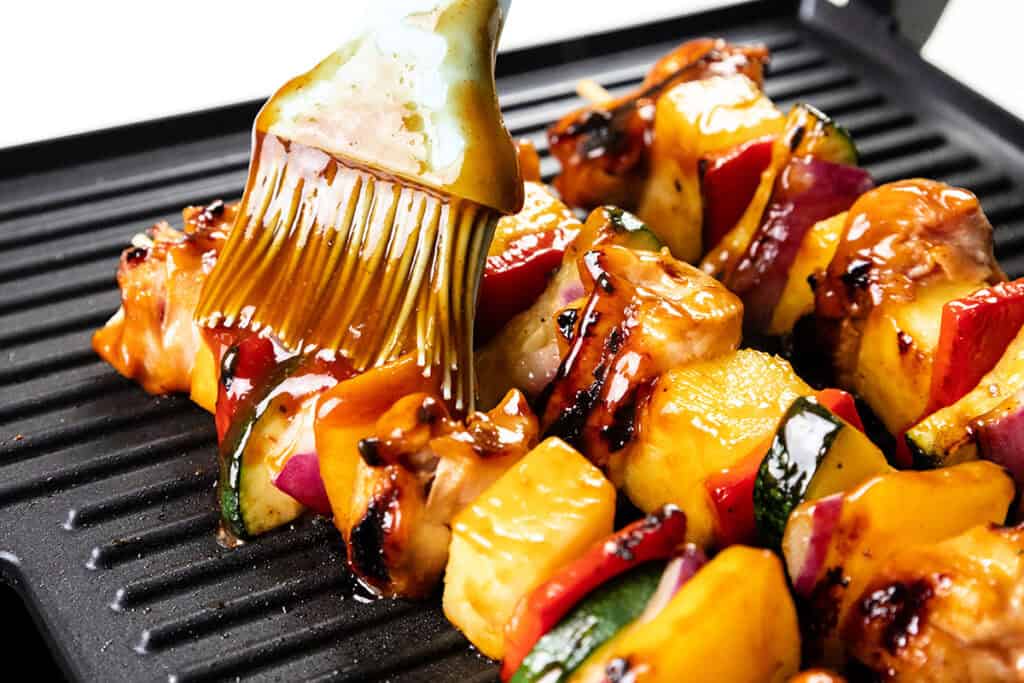 A close-up of BBQ chicken and vegetable skewers on a grill being brushed with a shiny glaze using a silicone brush. The skewers include pineapple, red onion, zucchini, and red bell pepper.