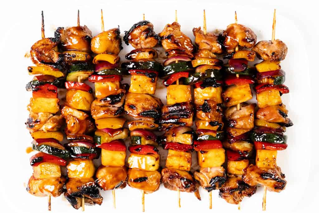 Grilled BBQ Chicken skewers with glazed chicken, pineapple, zucchini, red onion, and red bell pepper are arranged side by side on a white plate. The vegetables and meat appear perfectly charred and caramelized.